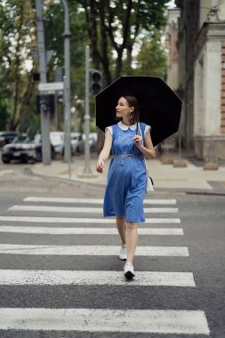 Mavi elbiseli, beyaz çantalı ve yaya geçidinden geçen şemsiyeli güzel bir kadın.