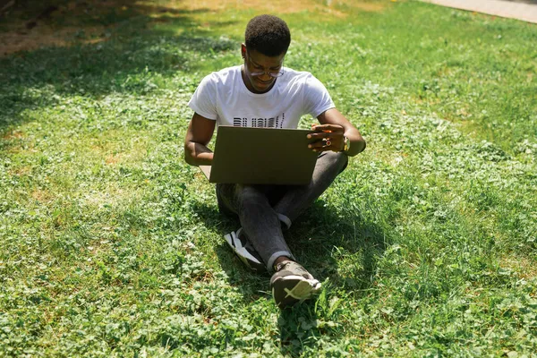 Yazın çimlerin üzerinde dizüstü bilgisayarı olan Afrikalı bir erkek çalışıyor.