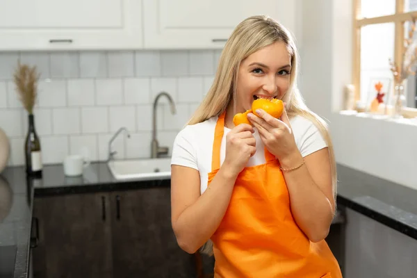 台所でオレンジを食べるエプロンの美しい女性の肖像画 — ストック写真
