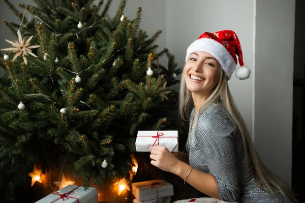 サンタ ハットの陽気な魅力的な若い女性クリスマスツリーの近くで手にプレゼントを保持 — ストック写真