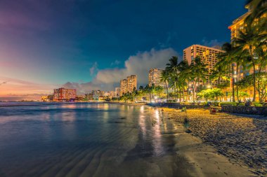 Honolulu, Hawaii 'de kum ve palmiye ağaçlarıyla Waikiki Sahili' nin gece manzarası. Gün batımı ışığı