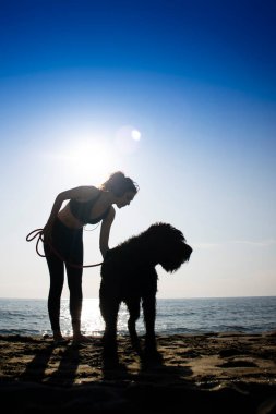 Kız, köpeğiyle birlikte deniz kenarında ışığa karşı yürüyor. 