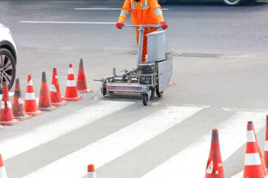Turuncu önlüklü bir yol işçisi yolda yaya geçidi işaretlerini yeniliyor ve işaretliyor..