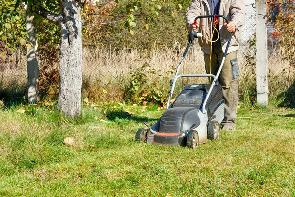 在阳光灿烂的秋天 一位园丁在自家后院照料着青草草坪 并用电动割草机割草 复制空间 — 图库照片