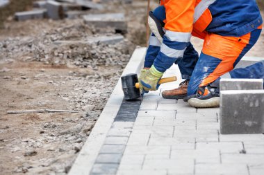Mavi ve turuncu tulumlu bir işçi kauçuk bir çekiçle kaldırıma çömelip kaldırım döşeyecek. Yakın çekim, seçici odak, kopyalama alanı.