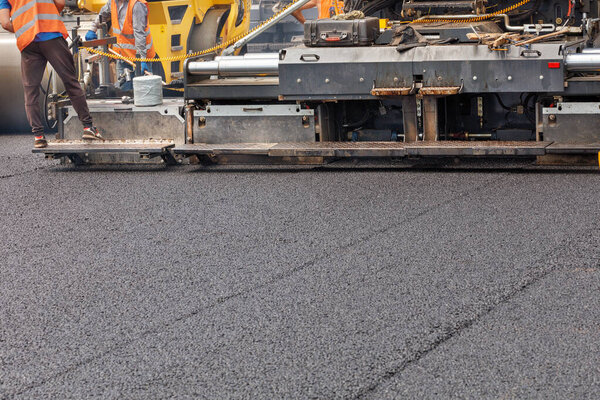 An industrial paver is laying a layer of new hot asphalt on the roadway. Workers control the evenness and height of the paving. Copy space.