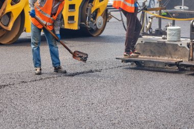 Yol işçileri, bir kürek, kaldırım ve yol silindiri ile yolun bir bölümünü onarıyor ve yola yeni bir sıcak asfalt tabakası seriyorlar. Boşluğu kopyala.