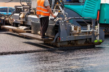 A road worker in an orange vest operates an asphalt paver and places hot fresh asphalt on a bitumen pavement while standing behind a control panel on a metal step on a summer day. Close-up, copy space. clipart