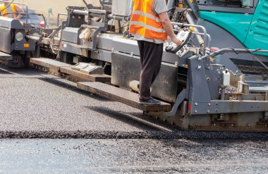 A road worker in an orange vest drives an asphalt paver and pours hot fresh asphalt onto a bitumen pavement on a summer day. Close-up, copy space.