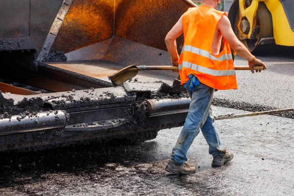 A worker in an orange vest scrapes fresh asphalt off a paver bucket with a shovel on a sunny summer day. Selective focus.