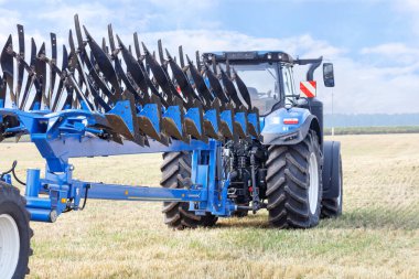 A metal multirow plow is attached to a tractor standing on an agricultural field and is ready for plowing after the harvest has been harvested. Copy space. Closeup.