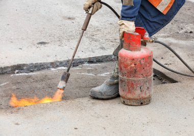 Yoldaki çukur tamiri. İşçi, bir gaz ocağı ve propan tankı kullanarak asfalt yüzeyini daha iyi ziftlenebilmek için ısıtıyor..