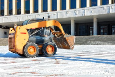 Açık bir kış gününde, manevra kabiliyeti yüksek bir memur idari binanın önündeki şehir meydanından kar yığınlarını temizler. Boşluğu kopyala.