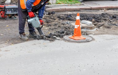 Yansıtıcı turuncu yelek giyen bir yol işçisi, benzin jeneratörüyle çalışan bir elektrikli matkap kullanarak kanalizasyon kapaklarının etrafındaki yolu onarıyor..