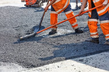 Turuncu tulumlu bir çalışma grubu, yolun bir bölümünü taze asfalt ile onarıp kürek ve metal seviyeleriyle düzleştiriyor. Boşluğu kopyala.