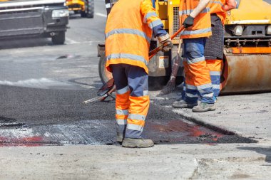 Turuncu tulumlu yol işçilerinden oluşan bir ekip, yolun küçük bir bölümünü onarmak için kürek, seviye, süpürge ve yol ekipmanları kullanarak yolun bir kısmını taze asfalt ile kapatıyor. Boşluğu kopyala.