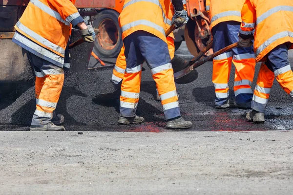一队身着橙色制服的道路工作人员用新鲜的沥青重新铺设了部分路面 并用铲子把路面铺好 然后用压路机压实 复制空间 — 图库照片