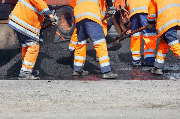 Turuncu üniformalı yol işçilerinden oluşan bir ekip yolun bir kısmını taze asfalt ile yeniden yüzeye çıkarıyor, küreklerle yayıyor, sonra da yol silindiriyle sıkıştırıyor. Boşluğu kopyala.