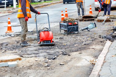Yolun turuncu trafik konileriyle çevrili kısmında, yol işçileri yıpranmış kanalizasyon kapaklarını yenileriyle değiştiriyor ve yüzeyi bir benzin rampasıyla dolduruyorlar..