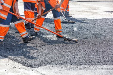 Turuncu yansıtıcı üniformalı yol işçileri güneşli bir yaz gününde yol kenarında metal seviyesiyle taze asfaltı düzleştiriyorlar. Boşluğu kopyala.