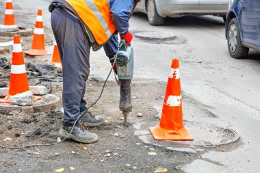 Bir yol işçisi, trafik konileriyle çevrili bir yol bölümünde elektrikli matkapla çalışıyor..