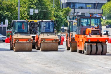 Turuncu endüstriyel yol silindirleri güneşli bir günde şehir caddesinde taze asfalt taşıyan kamyonların gelmesini bekliyor..