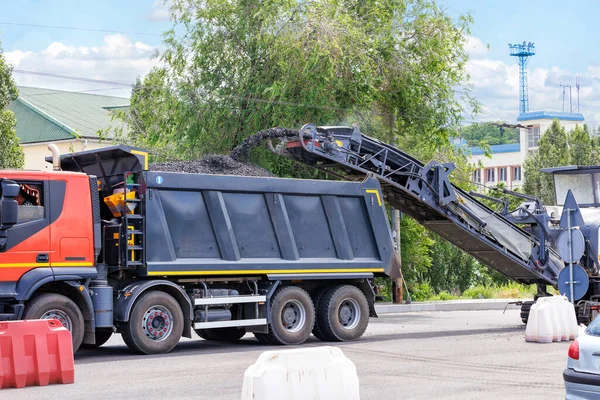 A road milling machine removes the top layer of old asphalt from the road and loads it onto a dump truck on a conveyor belt on a summer day. Copy space.