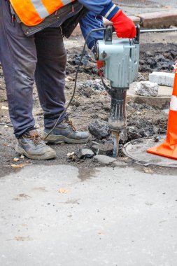 A road worker in a reflective orange vest is repairing a stretch of road around a sewer manhole using an electric jackhammer. Copy space, vertical image.