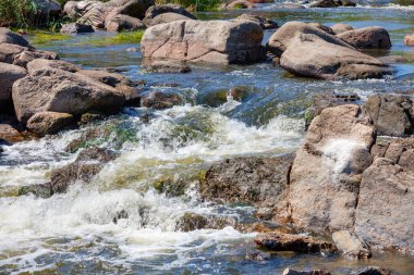 A blue stream of water seethes among rocky stone boulders on a sunny summer day.