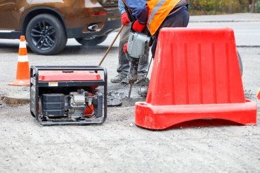 Road workers on a section of the road fenced with a red road shield are repairing the roadway with an electric jackhammer using a gasoline generator. Selective focus, copy space.