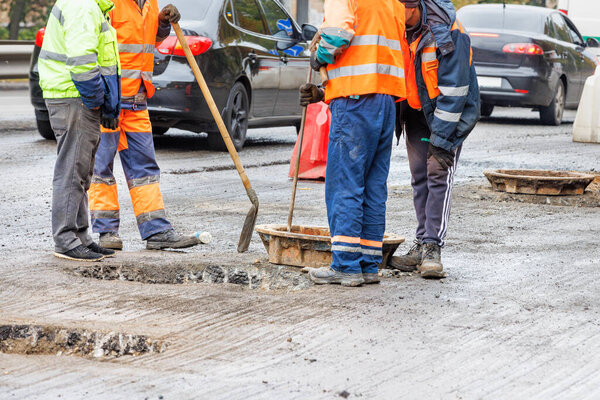 A group of road workers in orange and green reflective overalls are repairing a network of old sewer manholes in the roadway against the backdrop of passing cars in blur. Copy space.