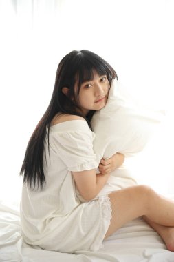 Güzel, genç Asyalı yastıkla yatakta oturuyor. Sabah ışığında dinleniyor ve beyaz odada dinleniyor.