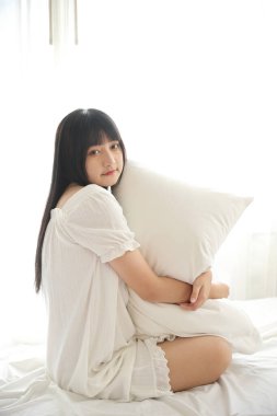 Güzel, genç Asyalı yastıkla yatakta oturuyor. Sabah ışığında dinleniyor ve beyaz odada dinleniyor.