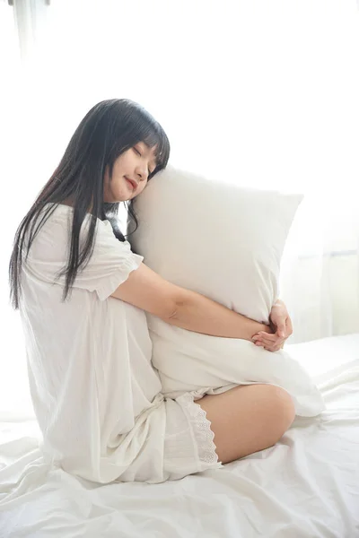 美しい若いアジア人は 白い部屋と朝の光の中でリラックスしたベッドの上に枕で座って — ストック写真