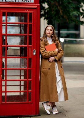 Kahverengi ceketli ve elinde turuncu defter, kırmızı telefon kulübesine yaslanmış beyaz elbiseli bir kadın.