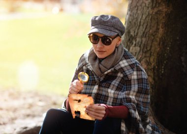 Sherlock Holmes gibi giyinmiş bir kadın parkta ağacın altında oturmuş elinde büyüteçle gazeteyi inceliyor.