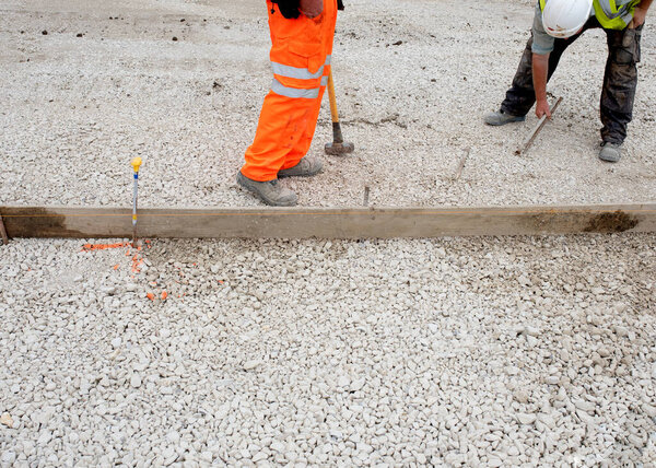 Groundworker in orange safety hi vis trousers fixing a timber along string line with steel pin to form a curb riser and a straight edge for tarmac road surface during new road construction