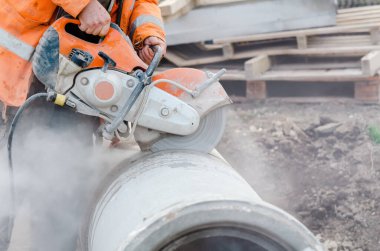 A worker at the construction site cutting a concrete drainage pipe with a petrol concrete saw. Builder covered in a hazardous dust cloud as safety procedure breached on construction site.