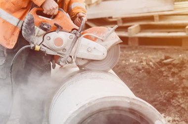 A worker at the construction site cutting a concrete drainage pipe with a petrol concrete saw. Builder covered in a hazardous dust cloud as safety procedure breached on construction site. clipart