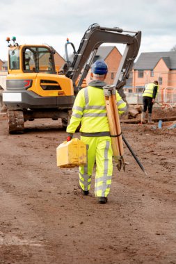 Site engineer wearing a bright hi-viz suit and safety boots walking on construction site carrying surveying equipment. Going to work on a daily basis concept