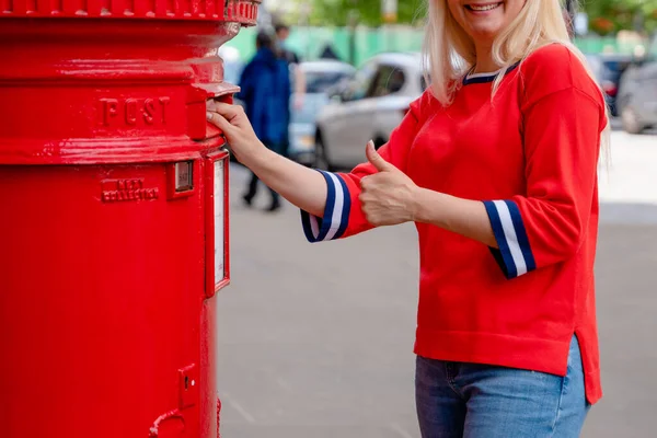 woman in red short posting letters in red post box in England