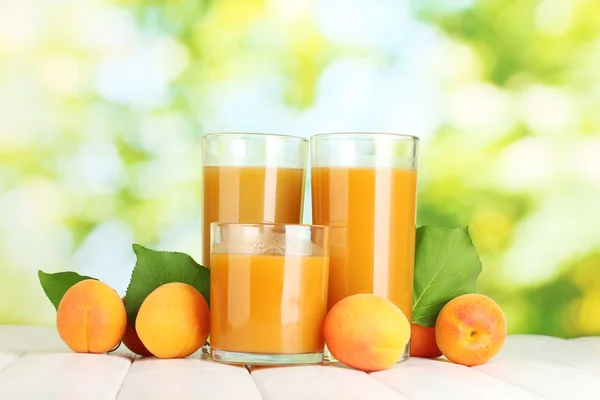 Glasses of apricot juice and fresh apricots on white wooden table on green — Stock Photo, Image