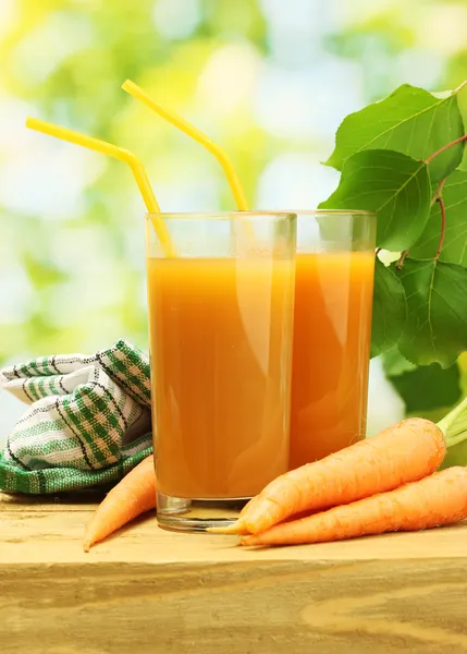Glasses of carrot juice and fresh carrots on wooden table on green backgrou — Stock Photo, Image
