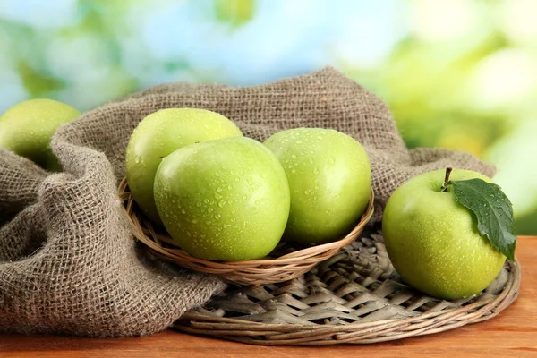 Ripe green apples in basket on burlap, on wooden table, on green background — Stock Photo, Image