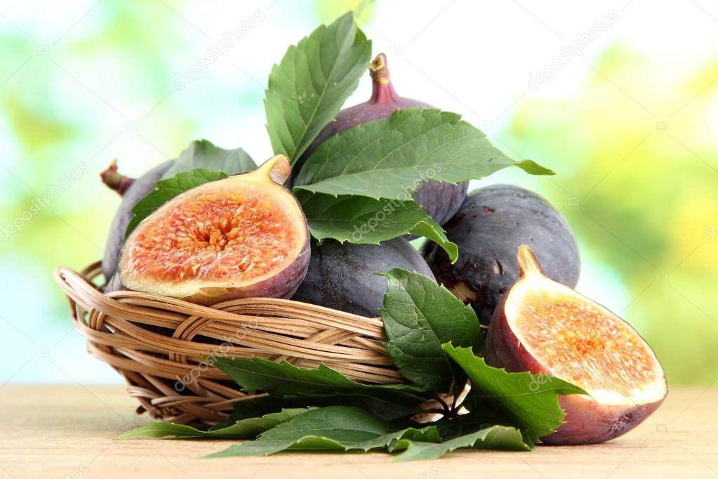 Ripe sweet figs with leaves in basket, on wooden table, on green background