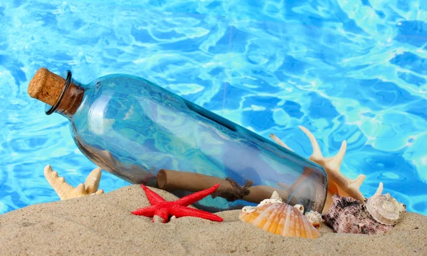 Glass bottle with note inside on sand, on blue sea background — Stock Photo, Image