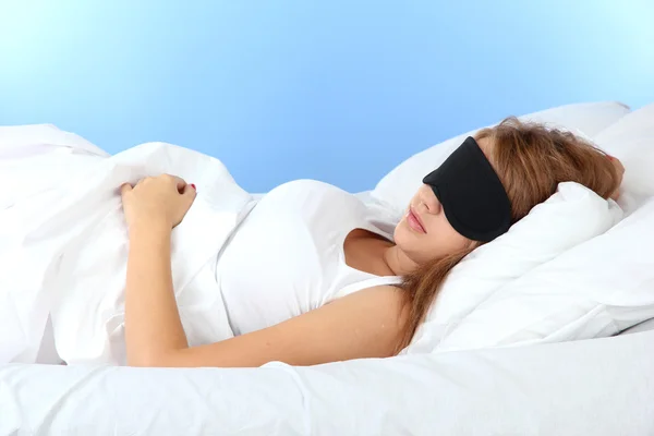 Jonge mooie vrouw slapen in bed met oog masker op blauwe achtergrond — Stockfoto