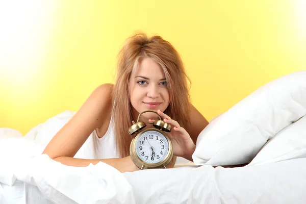 Young beautiful woman lying on bed with alarm clock on yellow background — Stock Photo, Image