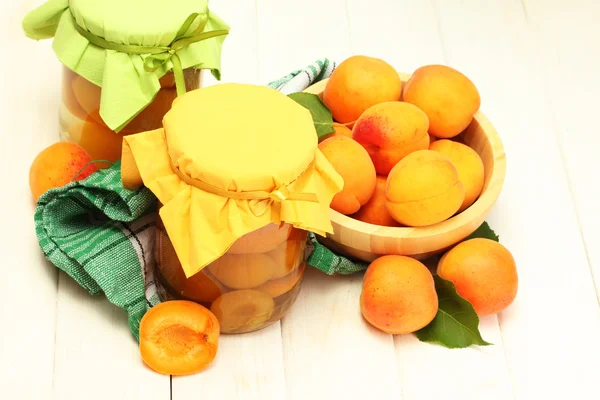 Canned apricots in a jars and ripe apricots in bowl on white wooden table — Stock Photo, Image