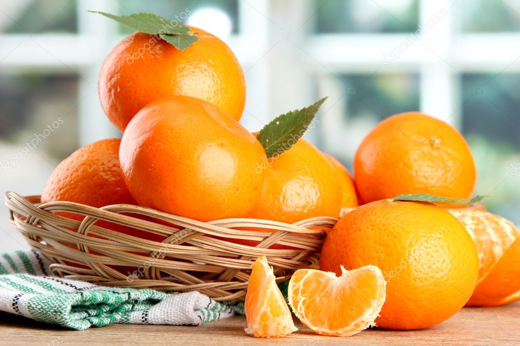 Tangerines with leaves in a beautiful basket, on wooden table on window background